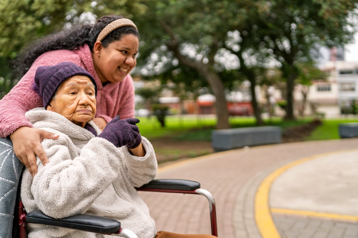 A young caregiver walking alongside a senior woman in a wheelchair on a path outdoors