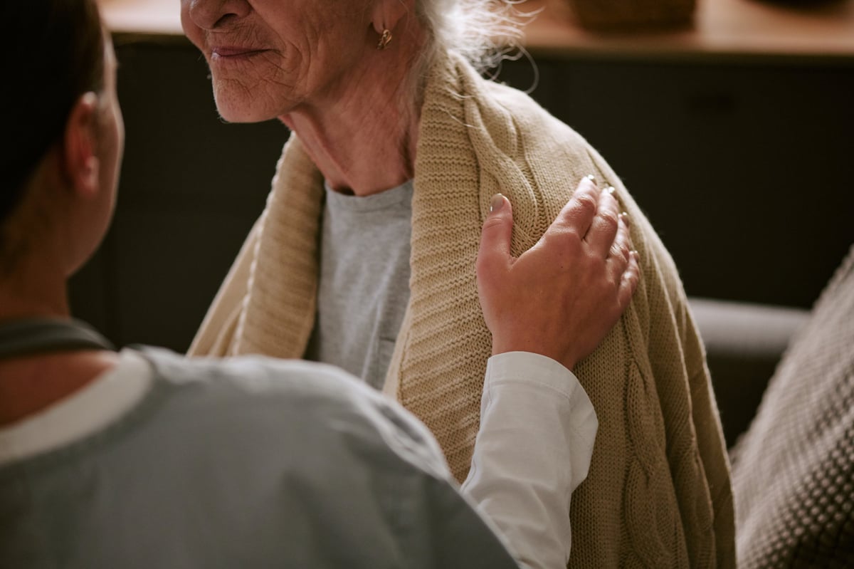 A young adult caregiver resting a hand on an older woman's shoulder in a warm, supportive gesture
